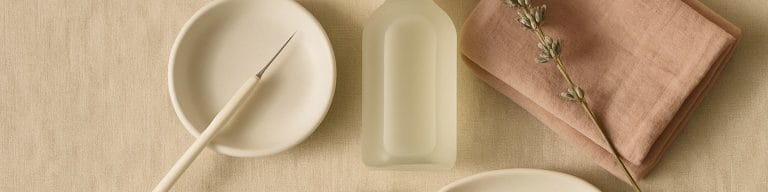 Editorial flat-lay of fine cosmetic skin tools, antiseptic bottle and magnifying lens on warm beige linen. Milia removal nea